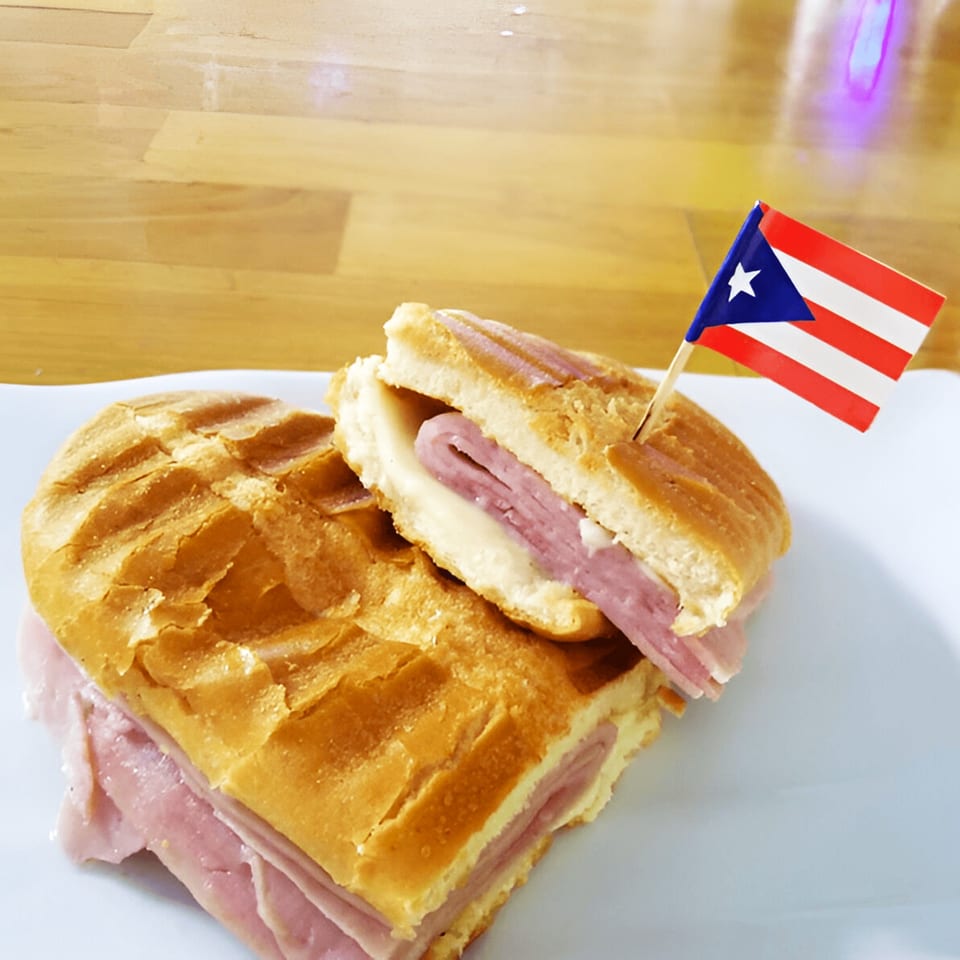 A Puerto Rican sandwich, cut in half, with ham and cheese, garnished with a small Puerto Rican flag.
