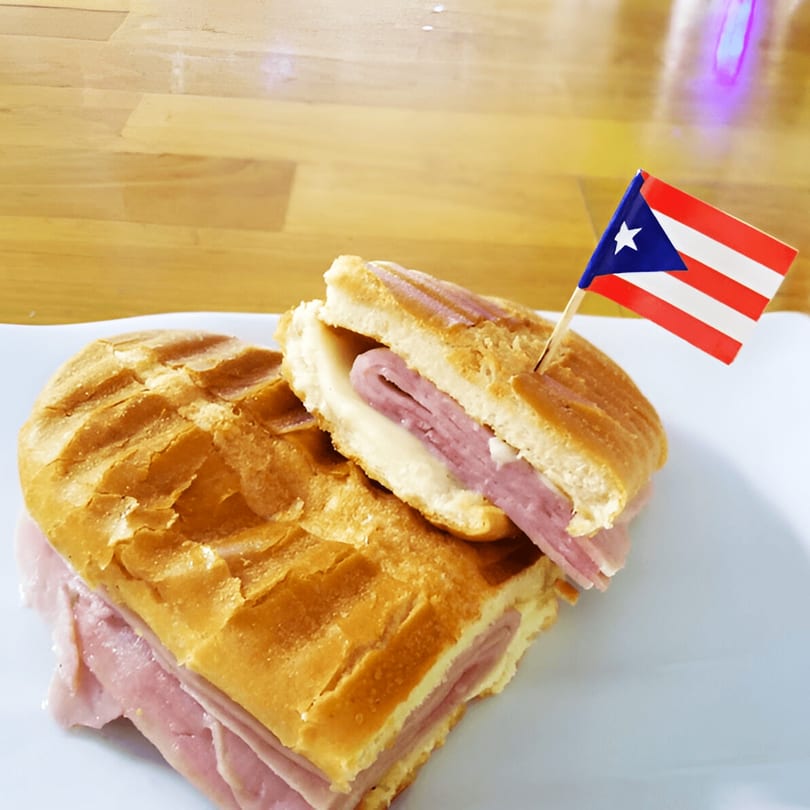 A Puerto Rican sandwich, cut in half, with ham and cheese, garnished with a small Puerto Rican flag.